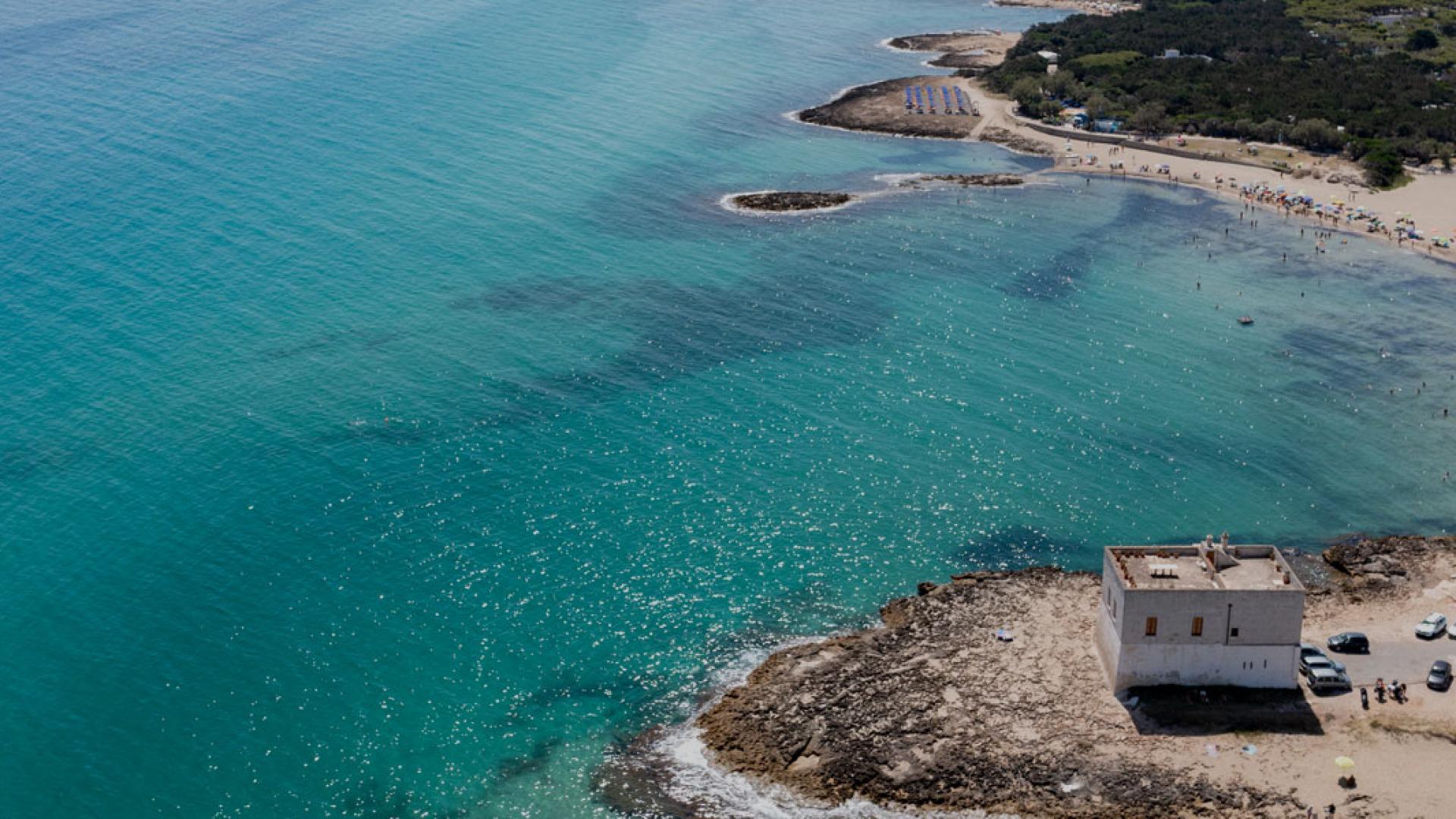 Spiaggia con mare cristallino, torre costiera e bagnanti sotto ombrelloni colorati.