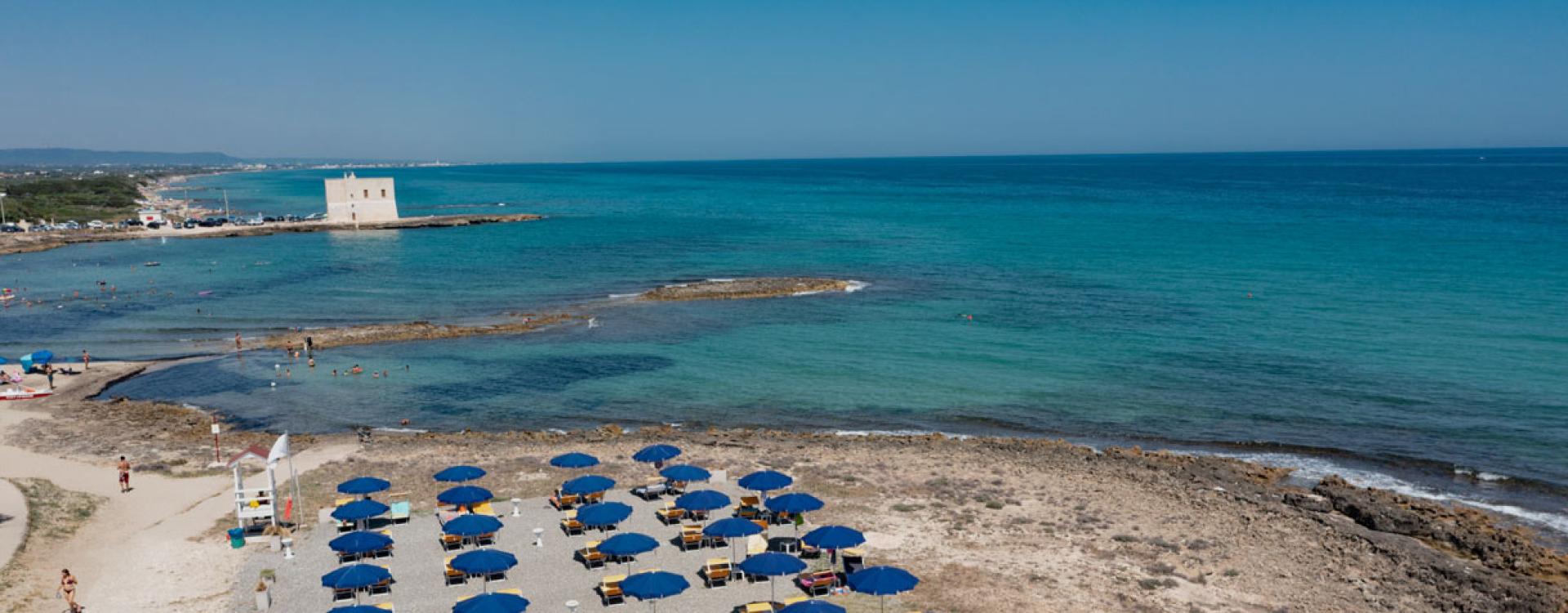 Spiaggia con ombrelloni blu e mare cristallino, torre sullo sfondo.