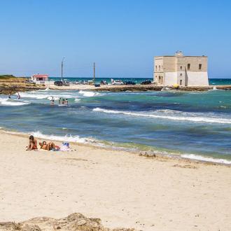 Spiaggia sabbiosa con bagnanti, mare mosso e edificio storico sullo sfondo.