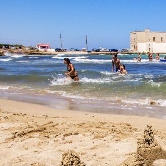 Spiaggia con bagnanti e torre costiera sullo sfondo, onde leggere e sabbia dorata.