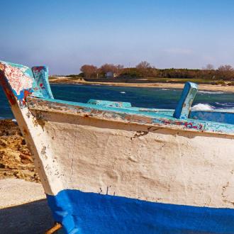 Vecchia barca blu e bianca sulla spiaggia rocciosa con mare e cielo sereno.
