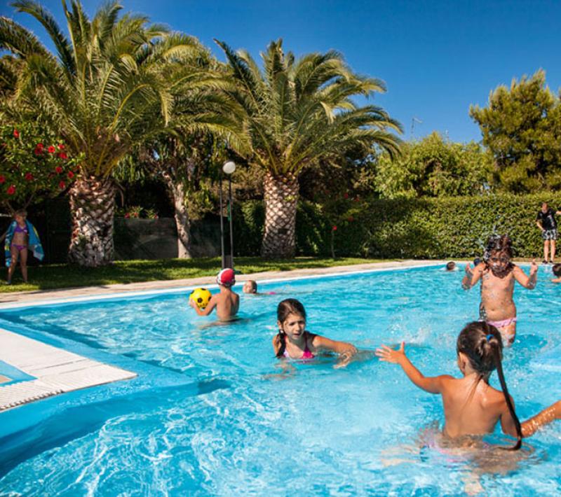 Bambini giocano in piscina sotto palme, giornata estiva e soleggiata.