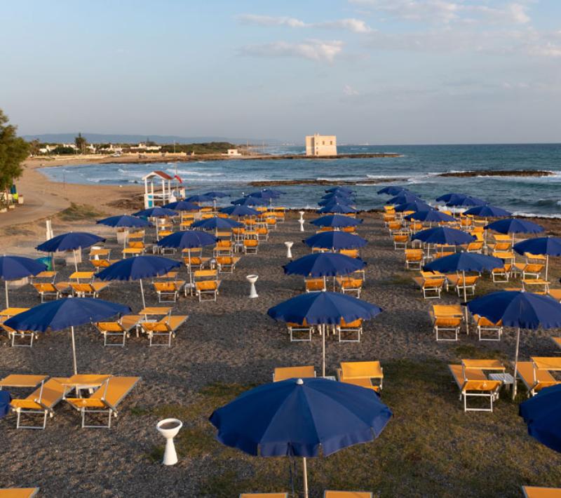 Spiaggia con ombrelloni blu e lettini gialli, mare calmo e torre sullo sfondo.