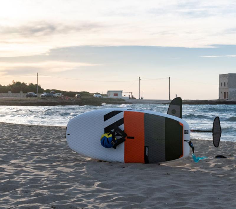 Tavola da surf sulla spiaggia al tramonto con mare mosso e edificio sullo sfondo.