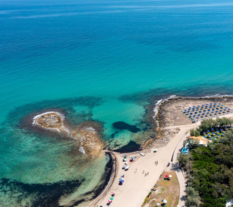Spiaggia con ombrelloni colorati e mare cristallino, circondata da scogli e vegetazione.